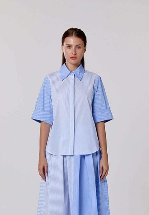 Femme portant une chemise à manches courtes rayée verticalement bleue et blanche et une jupe assortie, debout devant un fond uni.