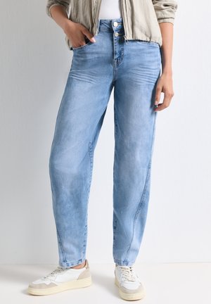 Frau trägt hellblaue Jeans mit hohem Bund, beige Jacke, weiße Sneakers, mit einer Hand in der Tasche vor hellem, einfarbigem Hintergrund.