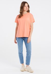 Perzikkleurig katoen T-shirt met korte mouwen, voorzien van een kleine witte palmbomenafbeelding op de linkerborst, gecombineerd met blauwe jeans en beige sneakers.