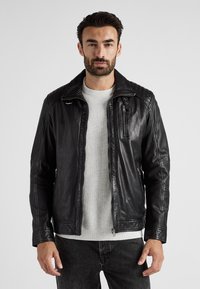 Svart läderjacka med ribbad axeldesign, hel dragkedja och sidoficka. Buren över en ljusgrå tröja.