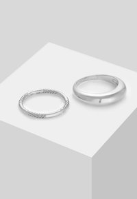 Två silverringar: en är en slät, bred bandring, och den andra har ett snorrigt repmönster med släta ändar. Båda är placerade på en ljus bakgrund.