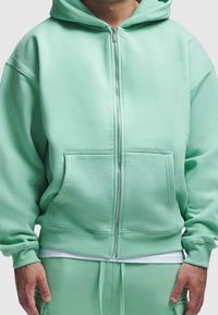 Homme portant un sweat à capuche zippé vert menthe et un pantalon de survêtement assorti, les mains détendues le long du corps.