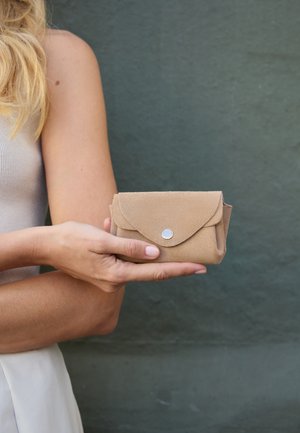 Woman holding a small beige suede clutch purse with a silver button, against a plain dark green wall background.