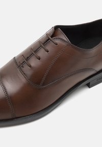 Pantof brown din piele, cu o finisare netedă, design cu șireturi și vârf rotund. Caracteristici include cusături subtile și un toc scăzut din cauciuc negru.