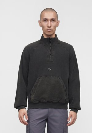 Mand med barberet hoved iført en sort kvartlynlås-pullover med en forlommer og grå cargobukser, stående mod en ensfarvet baggrund.