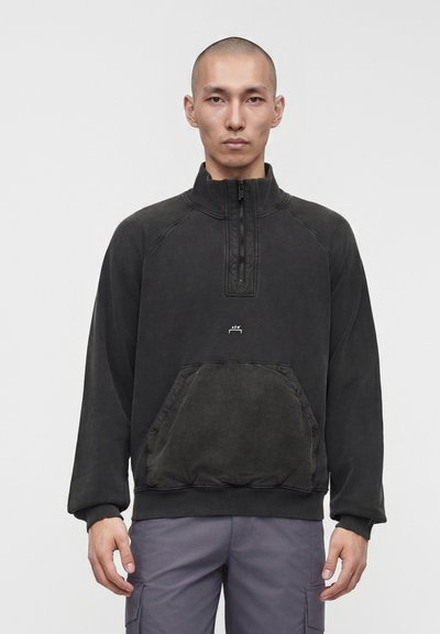 Homme à la tête rasée portant un pull noir à fermeture éclair quart de zip avec une poche avant et un pantalon cargo gris, debout devant un fond uni.