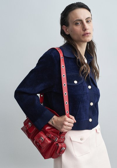 Woman in dark blue buttoned jacket and light skirt holding a red leather bag with buckle details over her shoulder.