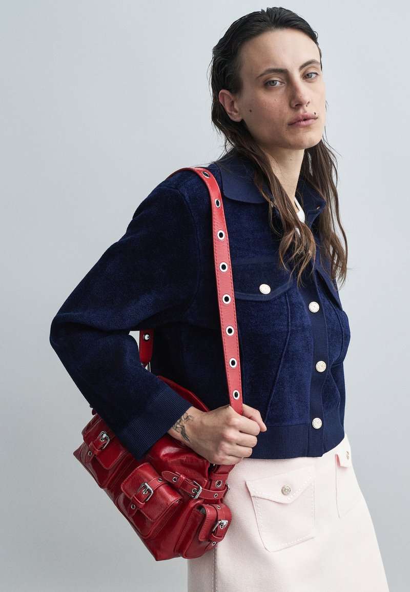 Mujer con chaqueta de botones azul oscuro y falda clara, sosteniendo un bolso de cuero rojo con detalles de hebilla sobre su hombro.