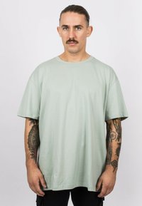 Mint green cotton t-shirt with short sleeves, round neck, and loose fit. Notable tattoos visible on the arms. Plain design, no patterns.