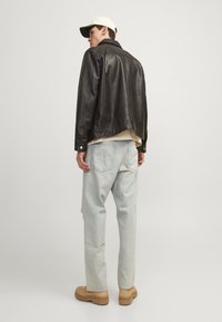 Svart skinnjakke med krage, lys beige skjorte under, lyseblå denimbukser og brune sko. Bakgrunnsvinkel, nøytral bakgrunn.