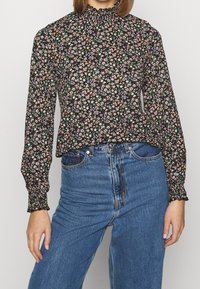 Zwarte blouse met bloemenprint, lange mouwen, geplooide halslijn en elastische manchetten. Gecombineerd met hoge blauwe denim jeans.
