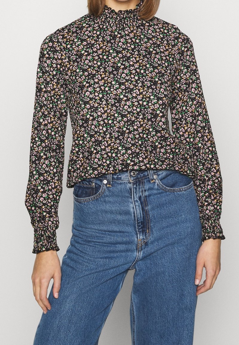 Zwarte blouse met bloemenprint, lange mouwen, geplooide halslijn en elastische manchetten. Gecombineerd met hoge blauwe denim jeans.