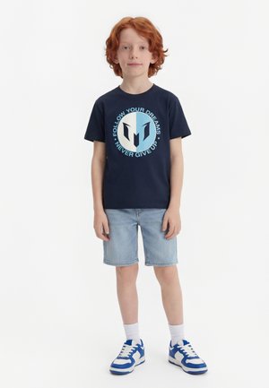 Un garçon aux cheveux roux est debout, portant un t-shirt bleu marine avec le texte "Suis tes rêves, n'abandonne jamais", un short en jean clair, des chaussettes blanches et des baskets bleu-blanc.
