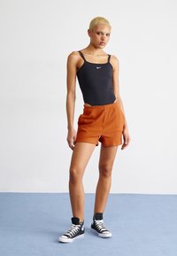Svart ärmlös bodysuit med Nike-logga, i kombination med strukturerade orange ribbade shorts. Modellen bär svarta högskurna sneakers och står poserad framför en blå bakgrund.