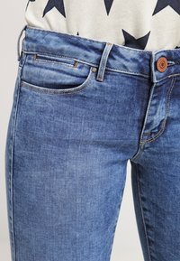 Blaue Denim-Jeans mit einer glatten Textur, die ein klassisches Fünf-Taschen-Design, Kupferknopf-Beschläge und kontrastierende Nähte aufweisen.
