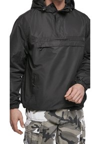 Svart lättviktsfleecejacka med en framficka, justerbar huva och elastiska ärmslut, i kombination med gråa och vita kamouflagebyxor.