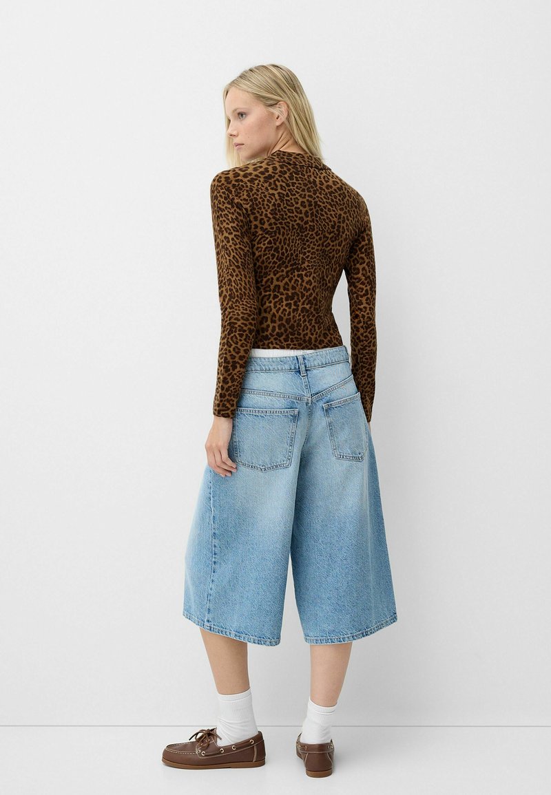 Langärmliges Oberteil mit braunem Leopardenmuster, kombiniert mit hellblauen Denim-Culottes, die locker sitzen und zwei Gesäßtaschen haben. Braune Schuhe mit weißen Socken.