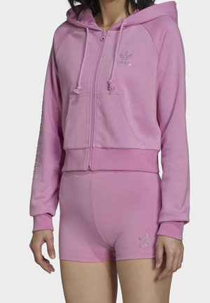 Femme portant un sweat à capuche rose zippé avec des cordons et un short rose assorti, avec de petits logos sur la poitrine et la jambe gauche.