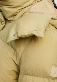 Jasnożółta kurtka puchowa o gładkiej fakturze, z pikowanymi sekcjami, napami oraz naszywką z logo z przodu.