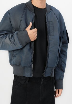 Mężczyzna w rozpiętej, ciemnoniebieskiej kurtce bombowej na czarnej koszulce i czarnych spodniach, poprawiający kołnierz kurtki lewą ręką.