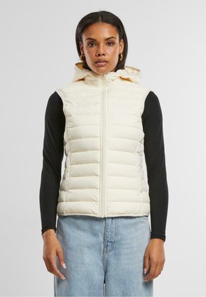 Cremfarbener Puffer-Weste mit Kapuze, mit horizontalem Steppmuster, Reißverschluss und kombiniert mit einem schwarzen Langarmoberteil und hellblauen Jeans.