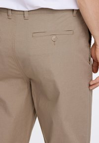 Beige lichtebroek van een textuurstof, met een achterzak met een knoopdetail en subtiele stiksel.