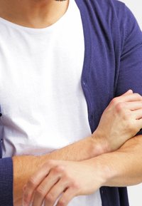 Mörkblå cardigan gjord av ett mjukt, strukturerat tyg, knäppt halvvägs, lagd över en enkel vit t-shirt, med synliga armar i kors.