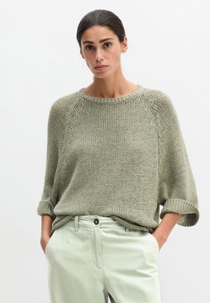 Kobieta w luźnym, oliwkowozielonym swetrze dzianinowym z podwiniętymi rękawami i jasnobeżowych spodniach, z rękami w kieszeniach, na prostym tle.