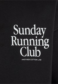 Tissu noir avec texte blanc indiquant « Sunday Running Club » et texte plus petit en dessous indiquant « Another Cotton Lab. »