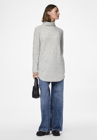 Sieviete pelēkā augstas apkakles džemperī, plašām zila džinsa biksēm, melnas krāsas apavos, turot nelielu melnu rokassomiņu, stāvot pret vienveidīgu baltu fonu.