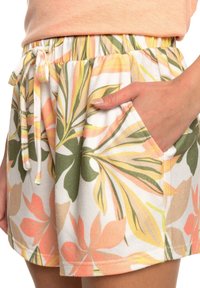 Main dans la poche portant un short blanc avec des motifs de feuilles tropicales vertes, jaunes et pêches, une taille élastique avec un cordon de serrage.