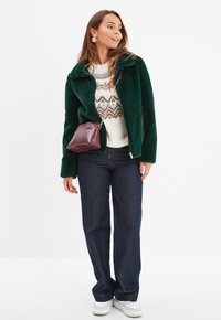 Grüne Kunstpelzjacke mit Reißverschluss, gemustertem Pullover, dunkelblauen Jeans und einer bordeauxfarbenen Umhängetasche mit strukturiertem Oberflächen, dazu weiße Sneakers.