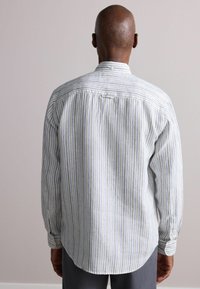 Long-sleeve striped shirt in light fabric, featuring blue and beige vertical stripes, curved hem, and buttoned cuffs. Back view shown.