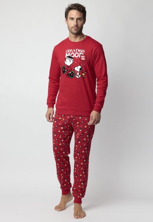 Hombre vestido con pijamas rojos de temática navideña con personajes de Peanuts y coronas navideñas, de pie descalzo sobre un fondo blanco.