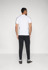 Witte sport T-shirt met korte mouwen en zwarte zijaccenten, gecombineerd met zwarte joggers en zwarte sportschoenen met een gele detail.