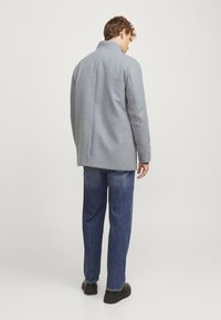 Abrigo de lana gris con un cuello alto, diseño ajustado y detalle de abertura lateral, llevado sobre unos pantalones de mezclilla azules y zapatos negros.