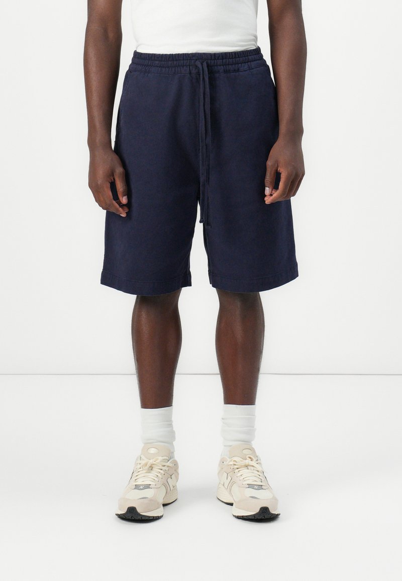 Pantalones cortos de algodón azul marino con cintura elástica y cordón. Presentan bolsillos laterales y un corte recto, combinados con calcetines blancos y zapatillas.