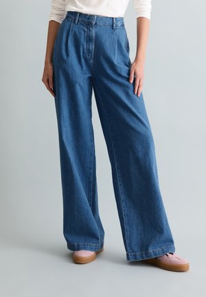 Osoba ubrana w wysokie, szerokie jeansy w kolorze niebieskim, z białą, długorękawową koszulą schowaną w spodniach oraz jasnoróżowymi butami na jednolitym tle.