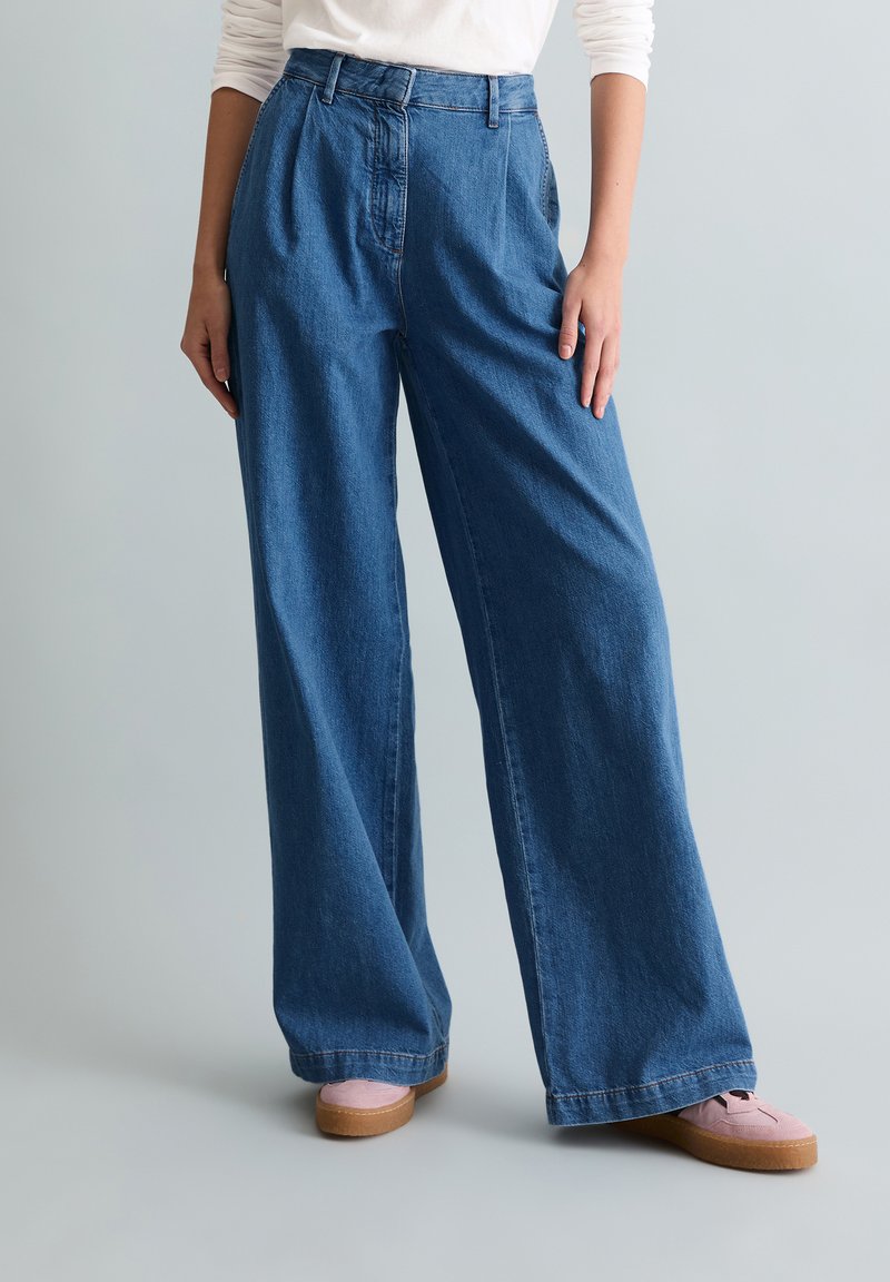 Άτομο που φοράει τζιν μπλε με ψηλή μέση και φαρδιά μπατζάκια, με λευκό μακρυμάνικο πουκάμισο κρυμμένο μέσα και ανοιχτό ροζ παπούτσια σε απλό φόντο.