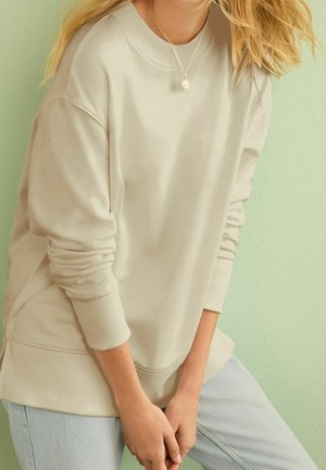 Osoba w beżowej bluzie i jasnoniebieskich dżinsach, lekko pochylona do przodu, z dłońmi splecionymi, oparta o jasnorozieloną ścianę.