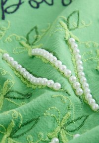 Collier de perles blanches reposant sur un tissu vert avec des feuilles et des vignes brodées dans diverses nuances de vert.
