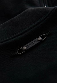 Czarna bluza z materiału z fakturowanym wykończeniem, z metalową metką z napisem „ambre” przymocowaną pętlą i zatrzaskiem.