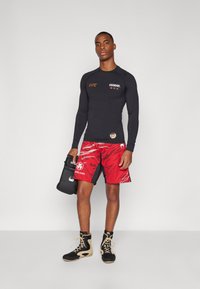 Haut à manches longues noir avec le logo UFC, shorts rouges avec un motif de rayures de tigre, gants de boxe noirs et chaussures de sport noir/or.