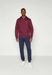 Bordo pulover s kapuco in z zadrgo ter sprednjimi žepi, kombiniran z mornarsko modrimi trenirkami. Bele superge dopolnjujejo izgled, ki prikazuje sproščen stil oblačenja.