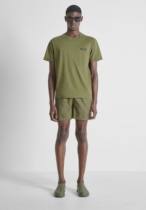 Hombre con camiseta verde oliva, pantalones cortos a juego, zapatos verdes sin cordones y gafas de sol negras, de pie frente a un fondo liso gris claro.