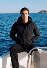 Homme portant une veste noire assis à l'avant d'un bateau avec les mains dans les poches, océan et île rocheuse visibles en arrière-plan.
