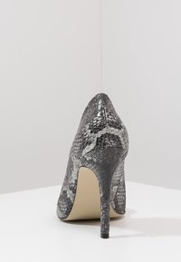 Högklackad sko med spetsig tå, tillverkad av strukturmaterial i grå ormskinnsdesign och en smal svart klack. Neutralfärgad sula.