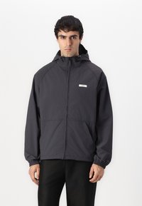 Schwarze leichte Jacke mit Reißverschluss, elastischen Bündchen, verstellbarem Kapuzen und kleinem Logo auf der linken Brustseite. Glatte Textur.