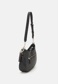 Sac à épaule noir avec sangle réglable, poche avant zippée et porte-clés étiqueté "1981" attaché à un anneau doré.