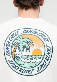 Άτομο που φοράει λευκό πουκάμισο με κυκλικό λογότυπο Santa Cruz, το οποίο περιλαμβάνει έναν φοίνικα, κύματα ωκεανού και πορτοκαλί ηλιοβασίλεμα.